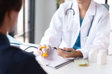 Bir hastaya ilacı açıklayan bir doktor. Doktor elinde reçeteli bir şişe tutuyor ve bir panoya yazı yazıyor. Tıbbi şişeler masada, tedavi hakkında detaylı bir tartışmayı gösteriyor..