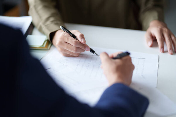 Close-up of two individuals discussing and pointing at a document on a desk. The hands are holding pens, indicating a review or collaboration on the content of the document.