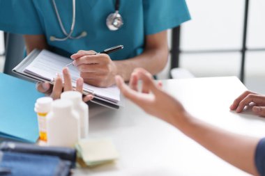 Mavi üniformalı bir doktor bir hastayla görüşürken not alıyor. Tıbbi şişeler ve belgeler masada.