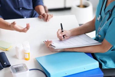 Mavi üniformalı bir doktor bir hastayla görüşürken not alıyor. Tıbbi şişeler ve belgeler masada.