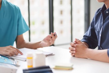 Mavi üniformalı bir doktor muayene sırasında bir hastayla tartışıyor. Tıbbi şişeler ve belgeler masada. Hasta elleri birbirine kenetlendi..