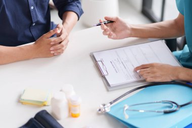 Mavi üniformalı bir doktor muayene sırasında bir hastayla tartışıyor. Tıbbi şişeler ve belgeler masada. Hasta elleri birbirine kenetlendi..