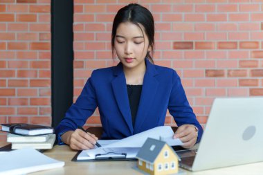 Mavi takım elbiseli bir iş kadını elinde kahve fincanı olan bir dizüstü bilgisayarla bir masada çalışıyor. Bir belgeye odaklanmış durumda..