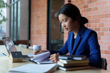 Mavi takım elbiseli bir iş kadını elinde kahve fincanı olan bir dizüstü bilgisayarla bir masada çalışıyor. Bir belgeye odaklanmış durumda..