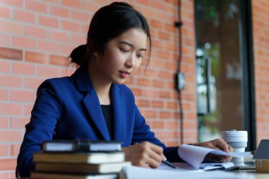 Mavi takım elbiseli bir iş kadını elinde kahve fincanı olan bir dizüstü bilgisayarla bir masada çalışıyor. Bir belgeye odaklanmış durumda..