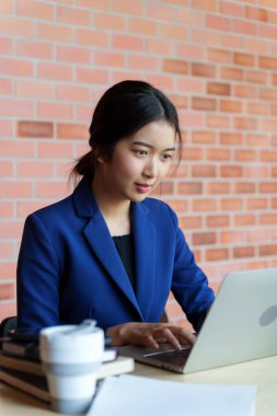 Mavi takım elbiseli bir ev satıcısı bir masada dizüstü bilgisayarı üzerinde çalışıyor. Arka planda tuğla bir duvar var. Masanın üzerinde, kitaplarla birlikte küçük bir model ev var.