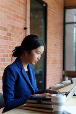 Mavi takım elbiseli bir ev satıcısı bir masada dizüstü bilgisayarı üzerinde çalışıyor. Arka planda tuğla bir duvar var. Masanın üzerinde, kitaplarla birlikte küçük bir model ev var.
