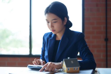Mavi takım elbiseli bir ev satıcısı masada hesap makinesiyle çalışıyor. Elinde bir kağıt parçası var ve hesaplamalarına odaklanmış görünüyor.. 