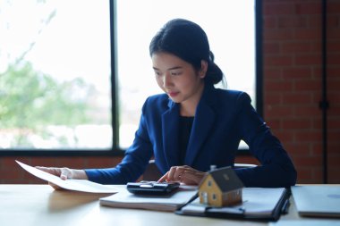 Mavi takım elbiseli bir ev satıcısı masada hesap makinesiyle çalışıyor. Elinde bir kağıt parçası var ve hesaplamalarına odaklanmış görünüyor.. 