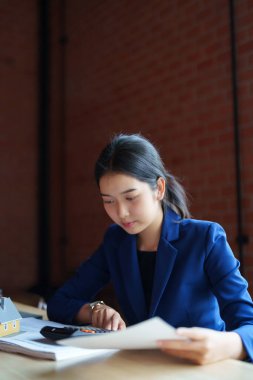 Mavi takım elbiseli bir ev satıcısı masada hesap makinesiyle çalışıyor. Elinde bir kağıt parçası var ve hesaplamalarına odaklanmış görünüyor.. 