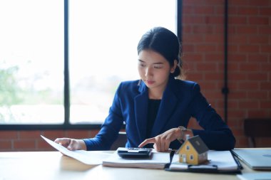 Mavi takım elbiseli bir ev satıcısı masada hesap makinesiyle çalışıyor. Elinde bir kağıt parçası var ve hesaplamalarına odaklanmış görünüyor.. 