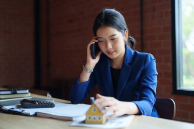 Mavi takım elbiseli bir ev satıcısı telefonla konuşurken model bir ev tutuyor. Gülümsüyor ve sohbet ediyor gibi görünüyor.. 