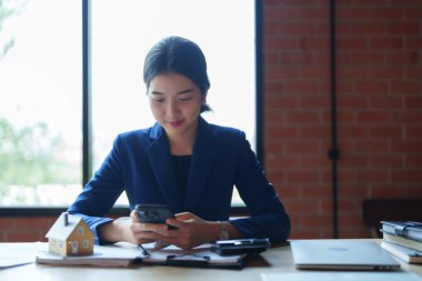 Mavi takım elbiseli bir ev satıcısı masada otururken telefonuna odaklanıyor. Büyük pencerelerden gelen parlak, doğal ışık çalışma alanını aydınlatıyor.