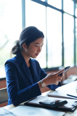 Mavi takım elbiseli bir ev satıcısı masada otururken telefonuna odaklanıyor. Büyük pencerelerden gelen parlak, doğal ışık çalışma alanını aydınlatıyor.