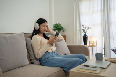 Genç Asyalı kadınlar eğlence sektöründe sörf yapmak ve kulaklıkla müzik dinlemek için akıllı telefonu kullanırken aynı zamanda rahat bir kanepede oturup evdeki yaşam tarzının tadını çıkarıyorlar..