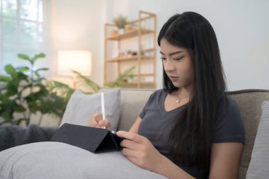 Genç Asyalı kadınlar proje pazarlama ve finans verileri hakkında iş raporları okurken ve kanepede oturup evde yaşam tarzının keyfini çıkararak rahatlamak için tablete notlar alırken.