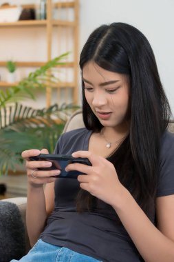 Genç Asyalı oyuncu kadınlar oyun oynamak için akıllı telefon kullanırken eğlence ve mutluluk ile rahat koltukta oturup evde yaşam tarzının tadını çıkararak zaman ve rahatlık geçiriyorlar..