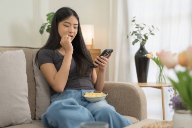 Genç Asyalı kadınlar sosyal medya eğlencesinde akıllı telefon kullanıyor ve kanepede oturup evde hayatın tadını çıkarırken mutlulukla abur cubur yiyorlar..