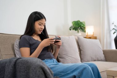 Genç Asyalı oyuncu kadınlar oyun oynamak için akıllı telefon kullanırken eğlence ve mutluluk ile rahat koltukta oturup evde yaşam tarzının tadını çıkararak zaman ve rahatlık geçiriyorlar..