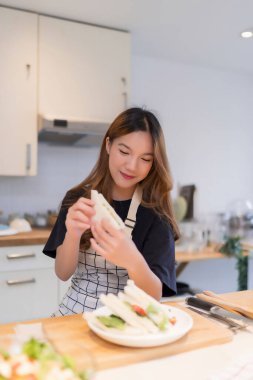 Genç Asyalı bir kadın lezzetli sandviçlerini yerken evde sağlıklı bir yaşam tarzı olan modern mutfakta taze sebze salatası ve ev yapımı atıştırmalıklar pişiriyor..