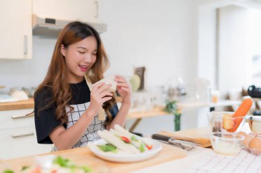 Genç Asyalı kadın kahvaltı yapıyor ve lezzetli sandviçler yiyor taze sebze pişirdikten sonra salata ve ev yapımı sağlıklı yemekler için ev yapımı atıştırmalıklar yapıyor..