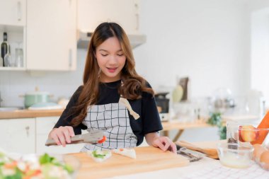 Genç Asyalı bir kadın, sağlıklı kahvaltı için lezzetli sandviç ve taze sebze salatası pişirmek için maşa kullanıyor. Ev yapımı sağlıklı yaşam tarzıyla modern mutfakta ev yapımı aperatif kesme tahtası hazırlamak için..