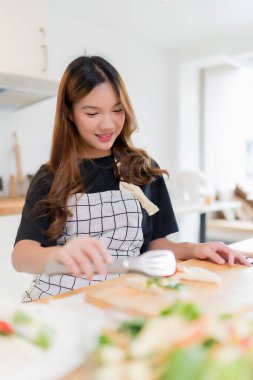 Genç Asyalı bir kadın, sağlıklı kahvaltı için lezzetli sandviç ve taze sebze salatası pişirmek için maşa kullanıyor. Ev yapımı sağlıklı yaşam tarzıyla modern mutfakta ev yapımı aperatif kesme tahtası hazırlamak için..