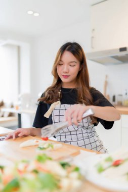 Genç Asyalı bir kadın, sağlıklı kahvaltı için lezzetli sandviç ve taze sebze salatası pişirmek için maşa kullanıyor. Ev yapımı sağlıklı yaşam tarzıyla modern mutfakta ev yapımı aperatif kesme tahtası hazırlamak için..