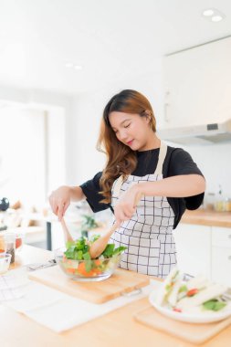 Genç Asyalı kadın taze salata sebzelerini kaseye karıştırmak için kaşık kullanıyor ve sağlıklı kahvaltı yemekleri için lezzetli sandviçler yapıyor ve modern mutfakta ev yapımı sağlıklı yaşam tarzıyla ev yapımı aperatifler yapıyor..