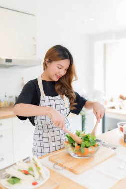 Genç Asyalı kadın taze salata sebzelerini kaseye karıştırmak için kaşık kullanıyor ve sağlıklı kahvaltı yemekleri için lezzetli sandviçler yapıyor ve modern mutfakta ev yapımı sağlıklı yaşam tarzıyla ev yapımı aperatifler yapıyor..