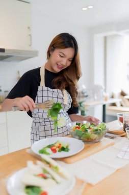 Genç Asyalı kadın, taze sebze salatasını kaseye karıştırdıktan ve modern mutfakta sağlıklı kahvaltı ve evde sağlıklı yaşam tarzını pişirdikten sonra maşa kullanarak tabağa sebze koyar..