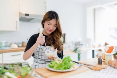 Genç Asyalı kadın tabağa mayonez koyarken kaşıkla salata koyuyor. Bu arada kâsede taze sebze salatası hazırlıyor ve evde sağlıklı yaşam tarzıyla modern mutfakta sağlıklı kahvaltı hazırlıyor..