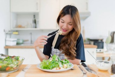 Genç Asyalı kadın sebze salatasıyla elma yiyor taze sebze salatasıyla salatayı sosla karıştırıp modern mutfakta sağlıklı kahvaltı hazırlıyor ve evde sağlıklı bir yaşam sürüyor..