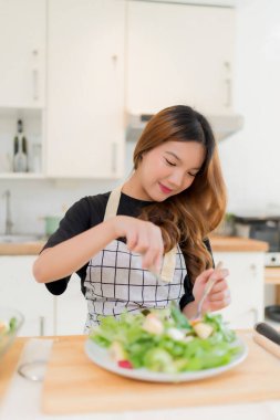 Genç Asyalı kadın taze sebze salatasıyla kaseye sarılmış salatayı karıştırdıktan sonra sebze yemekten hoşlanıyor ve modern mutfakta sağlıklı bir kahvaltı hazırlıyor ve evde sağlıklı bir yaşam sürüyor..