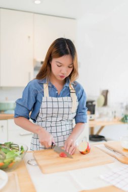 Genç Asyalı bir kadın bıçakla tahtayı keserken elma dilimlerken kâsede taze sebze salatası ve evde sağlıklı yaşam tarzıyla modern mutfakta sağlıklı kahvaltı hazırlıyor..