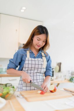 Genç Asyalı bir kadın bıçakla tahtayı keserken elma dilimlerken kâsede taze sebze salatası ve evde sağlıklı yaşam tarzıyla modern mutfakta sağlıklı kahvaltı hazırlıyor..