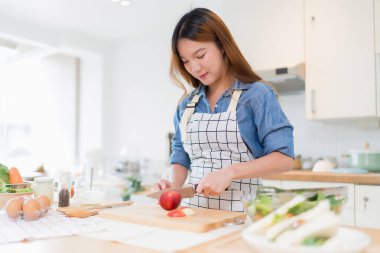 Genç Asyalı bir kadın bıçakla tahtayı keserken elma dilimlerken kâsede taze sebze salatası ve evde sağlıklı yaşam tarzıyla modern mutfakta sağlıklı kahvaltı hazırlıyor..