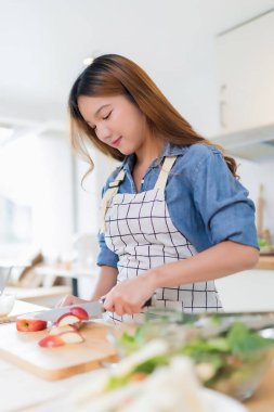 Genç Asyalı bir kadın bıçakla tahtayı keserken elma dilimlerken kâsede taze sebze salatası ve evde sağlıklı yaşam tarzıyla modern mutfakta sağlıklı kahvaltı hazırlıyor..