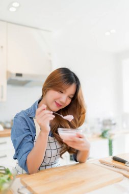 Genç Asyalı kadın taze sebze salatası hazırladıktan ve modern mutfakta sağlıklı yaşam tarzıyla sağlıklı kahvaltı yaptıktan sonra tatlı kaşığıyla lezzetli yoğurt yemeye kahvaltı ediyor..