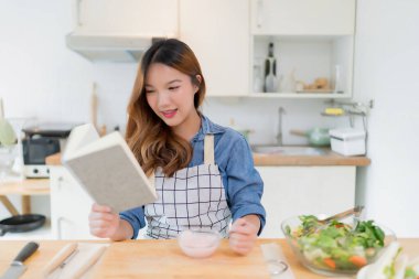 Genç Asyalı bir kadın lezzetli yoğurt yerken ve kitap üzerinde yemek tarifleri okurken aynı zamanda taze sebze salatası hazırlarken ve evde sağlıklı yaşam tarzıyla modern mutfakta sağlıklı kahvaltı hazırlarken..