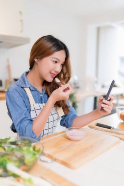 Yoğurt yiyen genç Asyalı bir kadın yemek tariflerinde akıllı telefon kullanırken aynı zamanda taze sebze salatası ve evde sağlıklı yaşam tarzıyla modern mutfakta sağlıklı kahvaltı hazırlıyor..