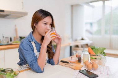 Genç Asyalı kadın elinde bir bardak portakal suyu tutarken ve taze meyve içerken sebze salatası hazırlarken ve evde sağlıklı yaşam tarzıyla modern mutfakta sağlıklı kahvaltı hazırlarken..