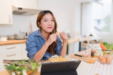 Genç Asyalı kadın atıştırmalık yiyor ve tablet kullanıyor. Taze sebzeler hazırlarken salata ve evde sağlıklı yaşam tarzıyla modern mutfakta sağlıklı kahvaltı hazırlıyor..