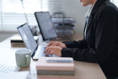 Siyah takım elbiseli profesyonel bir kadın ofis masasında oturuyor ve ofis malzemeleri ve belgelerle çevrili bir dizüstü bilgisayarla işine odaklanıyor..