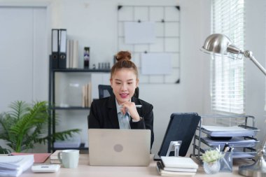 Siyah takım elbiseli profesyonel bir kadın masasında oturuyor. İşine yoğunlaşıyor. Etrafı dosyalar ve ofis malzemeleriyle çevrili..