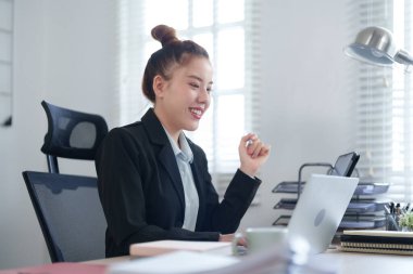 Siyah takım elbiseli profesyonel bir kadın masasında oturuyor. İşine yoğunlaşıyor. Etrafı dosyalar ve ofis malzemeleriyle çevrili..