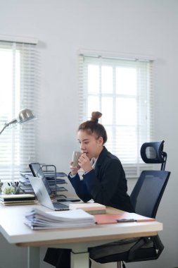 Siyah takım elbiseli kendine güvenen bir iş kadını masasında oturuyor, bilgisayarında çalışırken gülümsüyor, modern ve profesyonel bir çalışma alanı sergiliyor..