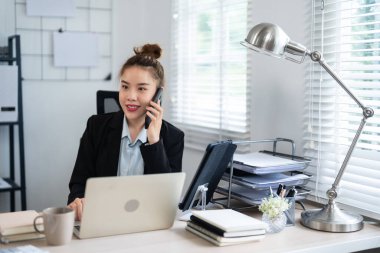 Siyah takım elbiseli bir iş kadını, masasında bilgisayarla çalışırken telefonla konuşuyor. Profesyonel ofis ortamında birden fazla görev ve verimliliği somutlaştırıyor..