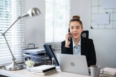 Siyah takım elbiseli bir iş kadını, masasında bilgisayarla çalışırken telefonla konuşuyor. Profesyonel ofis ortamında birden fazla görev ve verimliliği somutlaştırıyor..