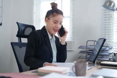 Siyah takım elbiseli bir iş kadını, masasında bilgisayarla çalışırken telefonla konuşuyor. Profesyonel ofis ortamında birden fazla görev ve verimliliği somutlaştırıyor..